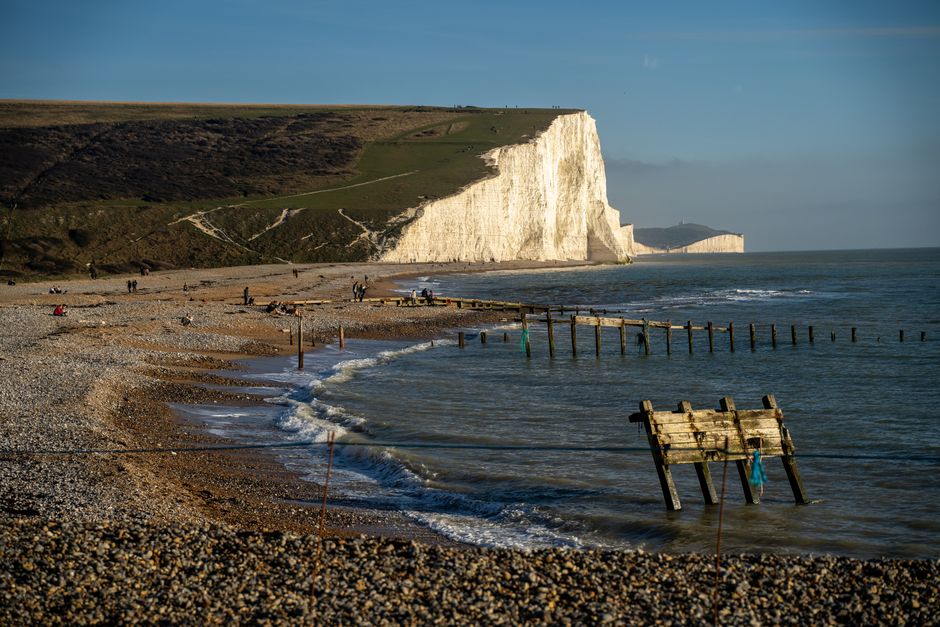 A panoramic view of a coastal landscape showing white chalk cliffs along a shoreline with a pebble beach at the base. In the foreground, there is a grassy area with patches of exposed soil and a small