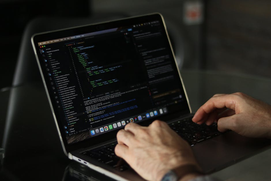Close-up of a person's hand with a wristwatch, typing on a silver laptop with a black keyboard. The laptop is placed on a wooden surface with visible grain and a warm brown finish. The screen displays
