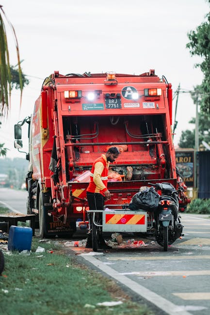 A sanitation worker wearing a yellow high-visibility vest and dark trousers is attending to a red rubbish collection truck parked at the roadside, with the truck's rear open to reveal its internal com